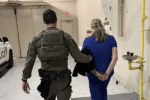 Law enforcement officer escorts a handcuffed woman in blue scrubs through a secure facility hallway.