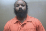 Mugshot-style image of a bearded man in an orange jail uniform against a plain gray wall.