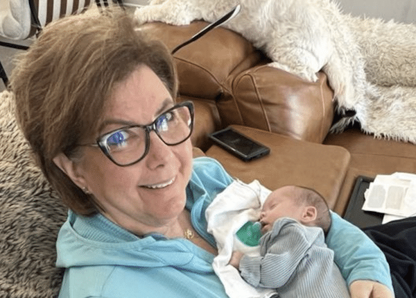 Woman wearing glasses smiles while holding a sleeping newborn on a couch.