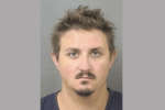 Mugshot-style portrait of a man with tousled hair and facial hair against a gray background.