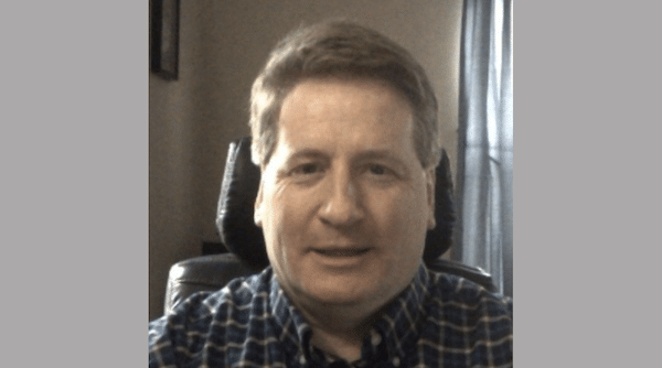 Webcam-style headshot of a middle-aged man wearing a blue plaid shirt and sitting in a black office chair.