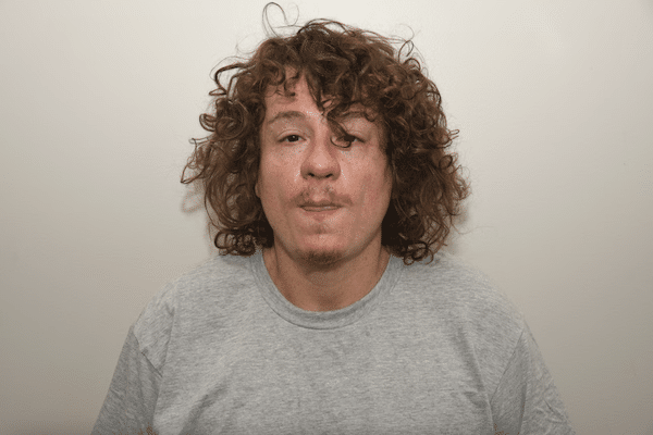 Head-and-shoulders image of a young man with curly red-brown hair wearing a gray T-shirt against a plain wall.