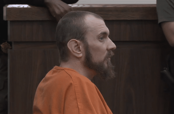 A bearded man in an orange jail uniform sits in profile inside a courtroom.
