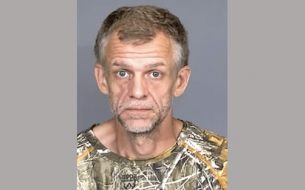Mugshot-style portrait shows a man in a camouflage shirt facing the camera against a gray background.