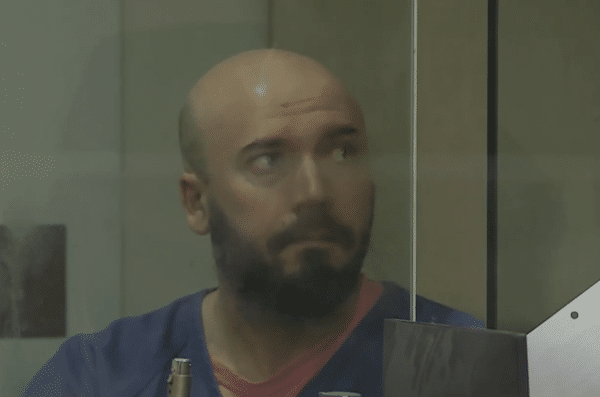 Bald bearded man sits behind glass while looking to the side.