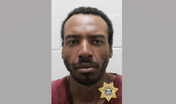 Booking-style portrait of a man in a maroon shirt with a sheriff’s badge graphic visible near his shoulder.