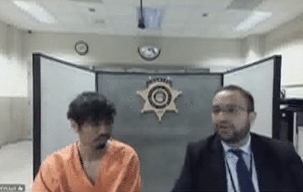 Man in an orange jail uniform sits beside an attorney during a remote courtroom appearance.