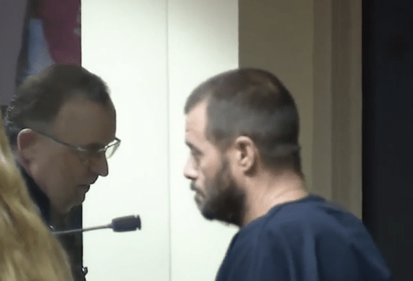 Side-profile courtroom image of a man with short hair and a beard wearing dark jail clothing.