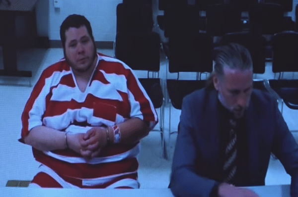 Man in a red-and-white striped jail uniform sits beside another man at a courtroom table.