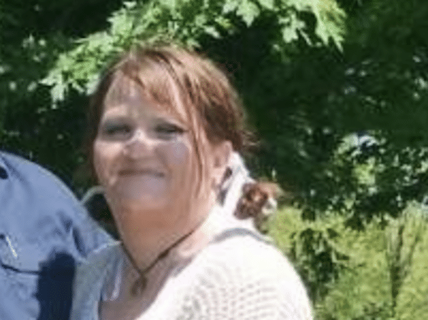 Outdoor photo of a smiling woman with reddish-brown hair standing near leafy green trees.