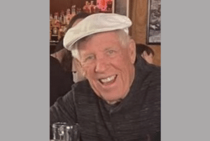 Photo of an older man smiling while wearing a light-colored flat cap in a restaurant or bar setting.