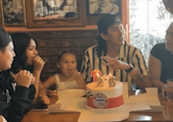 Family and friends sit around a table during a birthday celebration with a lit cake in front of them.