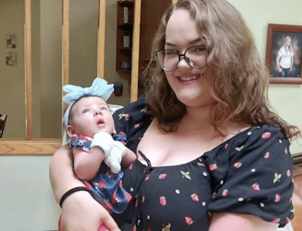 Woman wearing glasses smiles while holding a baby dressed in a blue outfit and headband.