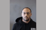 Portrait of a man in a black hoodie seated against a plain gray background.