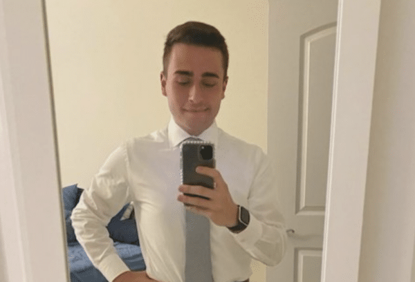 Man in a white dress shirt and gray tie taking a mirror selfie in a bedroom.
