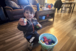 Young child smiling while holding up a small item beside a bucket filled with colorful plastic eggs in a living room.