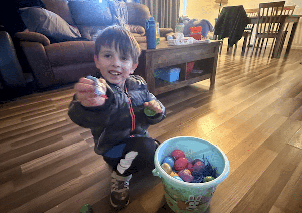 Young child smiling while holding up a small item beside a bucket filled with colorful plastic eggs in a living room.