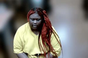 Woman with long red braids wearing a yellow shirt stands against a blurred background.