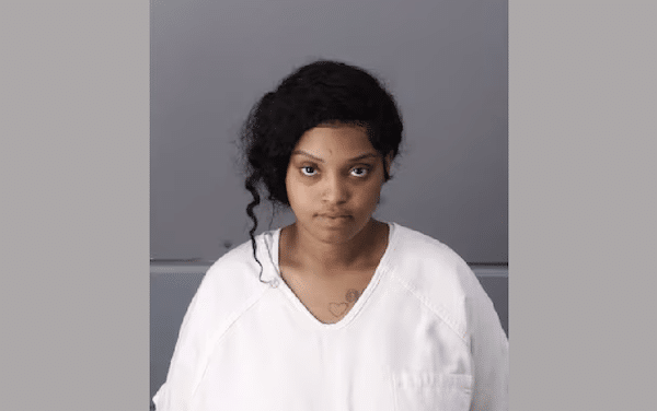 Booking-style portrait of a woman in a white jail uniform looking directly at the camera.
