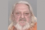 Mugshot-style portrait of an older man with long white hair and a full white beard wearing an orange shirt.