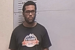 Mugshot-style image of a man wearing glasses and a black T-shirt, standing before a height chart.