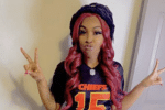 A woman with long red hair wears a Kansas City Chiefs jersey and holds up peace signs with both hands.