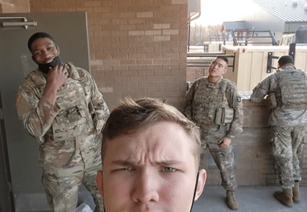 Four service members in camouflage uniforms stand outside a brick building, with one taking a close-up selfie in the foreground.