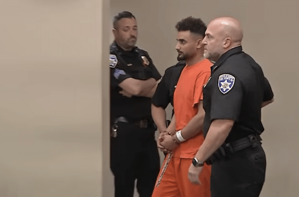 Man in an orange jail uniform stands in a courtroom beside law enforcement officers.