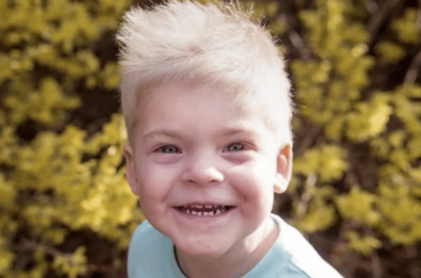 Smiling young boy with bright blond hair poses outdoors in front of blurred green foliage.