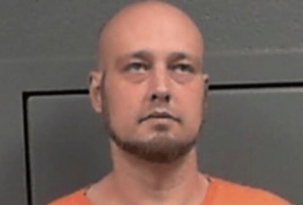 Close-up portrait of a bald man with a trimmed beard wearing an orange jail uniform against a gray background.