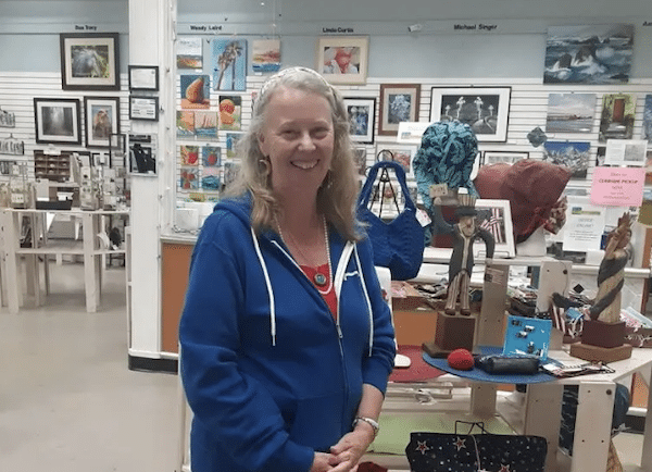 Smiling woman with gray-blond hair stands inside an art gallery or gift shop, wearing a blue zip-up hoodie among displayed artwork and crafts.