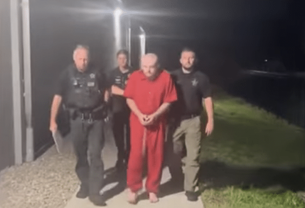 Three officers escort a barefoot man in red clothing along a walkway outside a building at night.
