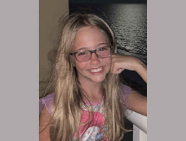 Smiling young girl with long blonde hair and glasses posing indoors with one hand resting near her face.