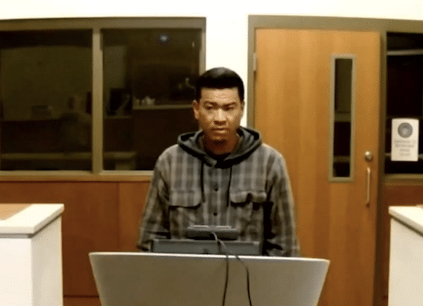 Man with short dark hair stands behind a lectern in what appears to be a courtroom or hearing room.
