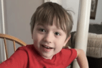 Young boy in a red T-shirt smiles toward the camera while seated indoors near a wooden chair.