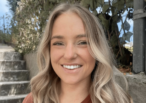 Close-up outdoor portrait of a smiling woman with long, wavy blonde hair, with concrete steps and greenery in the background.