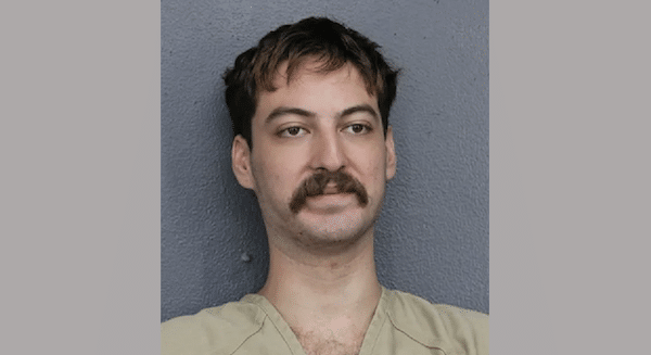 Mugshot-style portrait of a man with short dark hair and a thick mustache wearing a tan jail uniform against a gray wall.