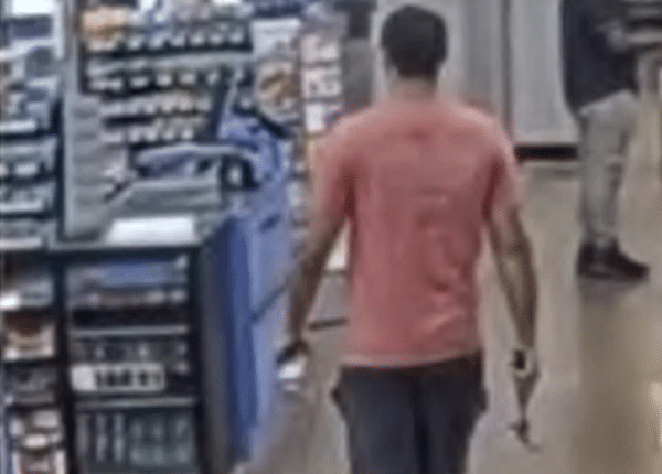 Blurry surveillance-style image of a man in a red shirt walking through a retail store aisle, seen from behind.
