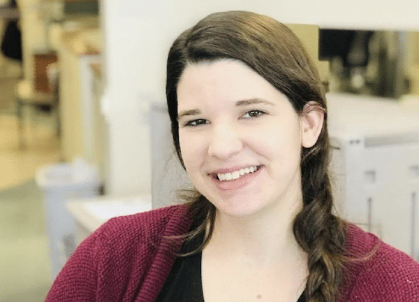 Smiling woman with brown hair in a side braid wears a burgundy cardigan in an indoor office or clinic setting.