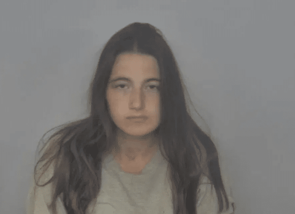 Mugshot-style photograph of a young woman with long dark hair wearing a gray shirt against a plain gray background.