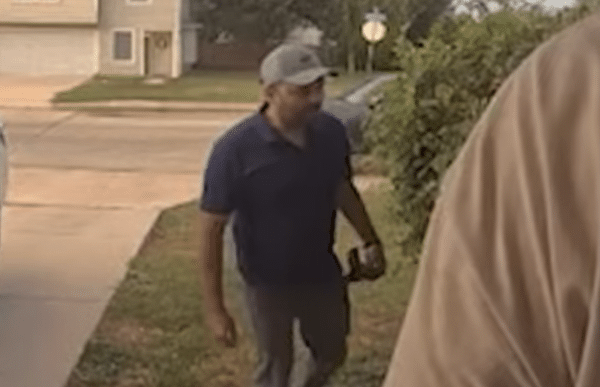 Man in a baseball cap and dark polo shirt walks along a grassy area beside a residential street.