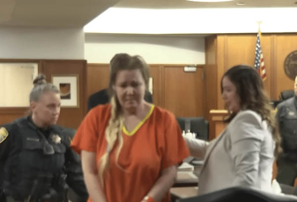 A woman in an orange jail uniform stands in a courtroom between a uniformed officer and another woman in professional attire.