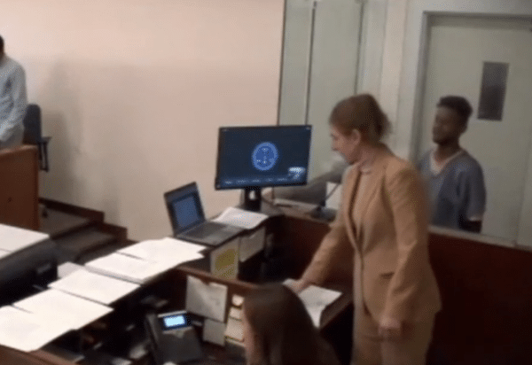 Courtroom scene showing a woman in a tan suit standing near a clerk’s desk while two men remain behind her near the wall.