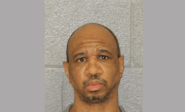 Mugshot-style portrait of a bald man with short facial hair looking directly at the camera against a gray block wall.