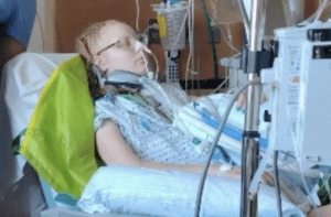 Hospital patient wearing a gown and neck brace lies in a bed surrounded by medical equipment and tubing.