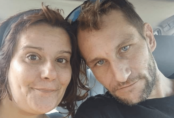 Close-up selfie of a woman with reddish-brown hair and a man with short hair and facial stubble sitting together inside a vehicle.