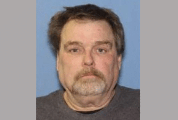 Mugshot-style portrait of a middle-aged white man with short brown hair, a mustache and goatee against a blue background.