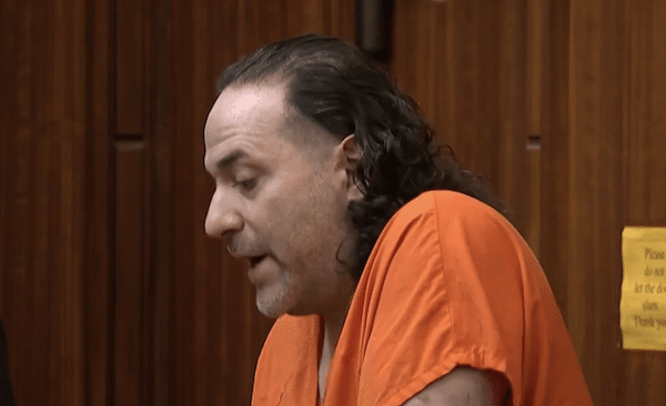 Courtroom image of a man with long dark hair and facial hair wearing an orange jail uniform while seated against wood-paneled walls.