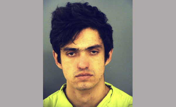 Young man with thick dark hair and light facial hair faces the camera against a plain gray background, wearing a bright green shirt.