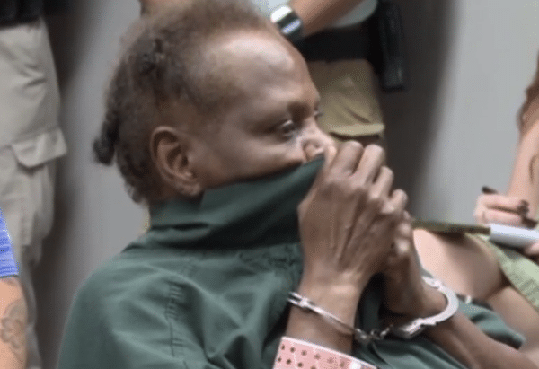 Man in a green shirt sits with his hands clasped near his face, wearing handcuffs as several people stand behind him.